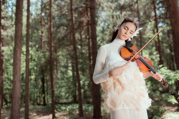 dromerige vrouw in witte zwaan kostuum staande op forest achtergrond, spelen op viool - Foto, afbeelding