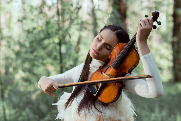 felnőtt nő fehér hattyú jelmez állva az erdő háttérben, hegedűre játszik - Fotó, kép