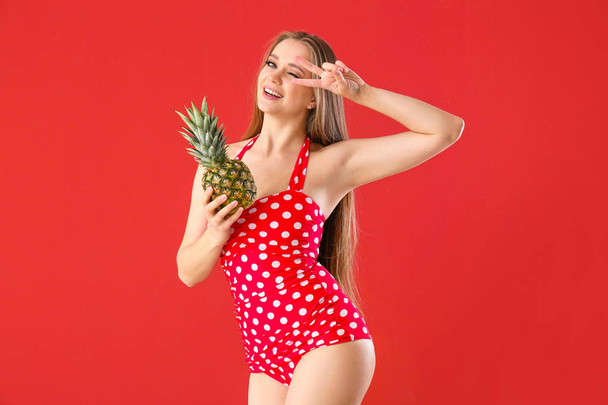 Beautiful young woman in swimming suit and with pineapple showing victory gesture on color background - Valokuva, kuva