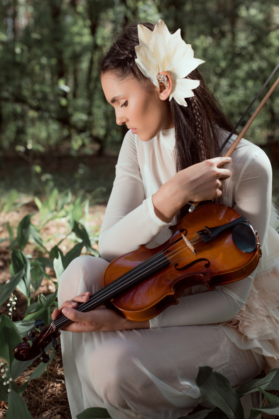 mujer joven en traje de cisne blanco sentada en el suelo con el fondo del bosque, mirando hacia otro lado, sosteniendo violín
 - Foto, imagen