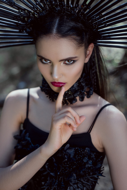 high angle view of young woman in witch costume with crown on head holding finger near lips, making shh sign, looking at camera - Photo, Image