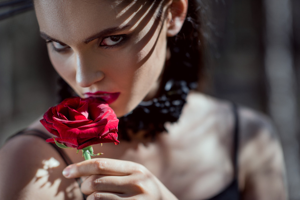 portrait of beautiful woman in black costume holding red rose in hand, looking at camera - Photo, Image