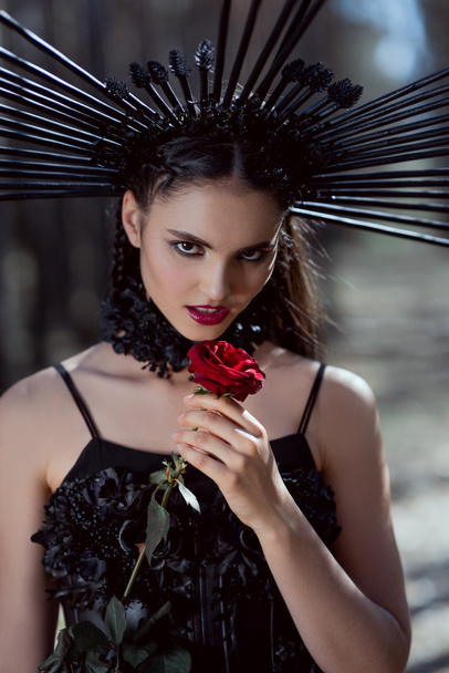 junge Frau im Hexenkostüm blickt in die Kamera und hält rote Rose in der Nähe des Gesichts - Foto, Bild