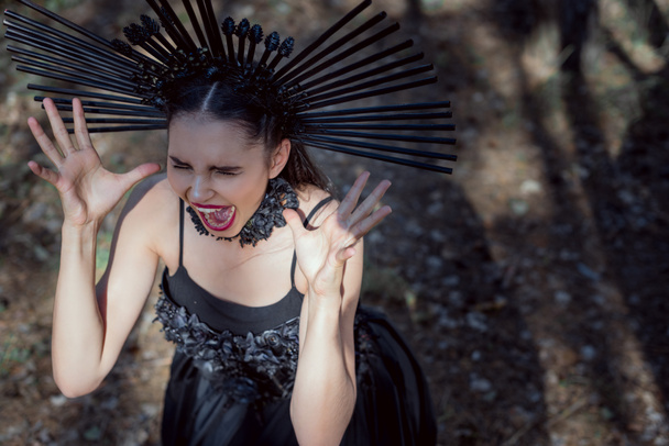 Высокий угол обзора сумасшедшей женщины в костюме ведьмы с короной на голове крича, закрывая глаза, держа руку возле лица
 - Фото, изображение