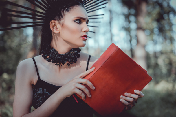 beautiful woman in witch costume standing on forest background, holding red book, looking away - Photo, Image
