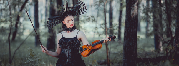 panoramic view of woman in witch costume with crown standing on forest background, holding violin - Photo, Image
