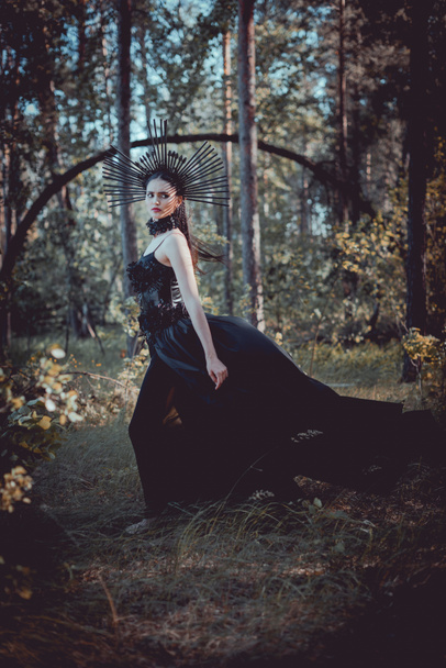πλευρική όψη της μελαχρινή γυναίκα με στολή μάγισσας στέκεται στο φόντο του δάσους, κοιτάζοντας μακριά - Φωτογραφία, εικόνα