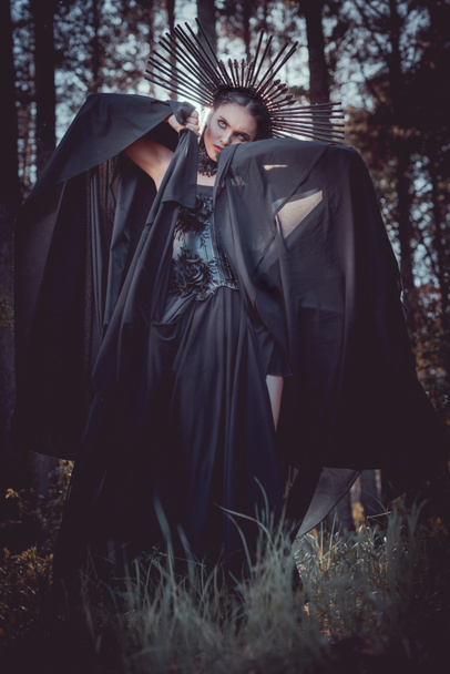 μελαχρινή γυναίκα με στολή μάγισσας και στέμμα στο κεφάλι κρατώντας τα χέρια με μαύρο ύφασμα κοντά στο πρόσωπο, κοιτάζοντας μακριά - Φωτογραφία, εικόνα