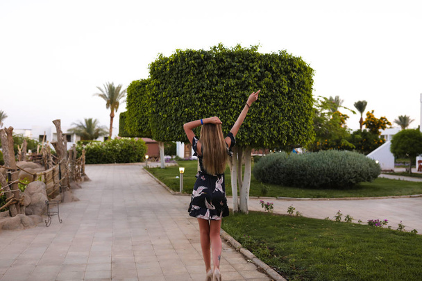 Modern black dress with birds pattern, light summer collection. High heel shoes, accessories. Young woman walking alone in a yard of the hotel, raised her arms. Long blonde hairstyle. Palm trees - Photo, Image