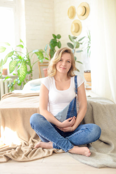 Beautiful pregnant woman with blond hair in a white T-shirt and blue jeans is sitting on a bed . Concept of happy motherhood, healthy lifestyle - Photo, Image