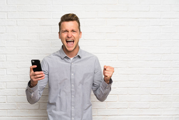 Blonde man over white brick wall with phone in victory position - Zdjęcie, obraz