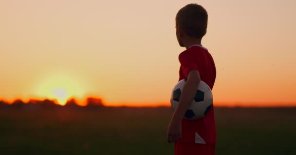 Een jongen met een voetbal kijkt naar de zonsopgang. - Video