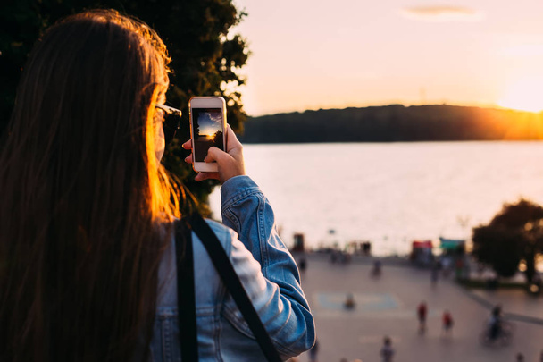 Το κορίτσι κρατά το κινητό της τηλέφωνο και φωτογραφίζει το ηλιοβασίλεμα - Φωτογραφία, εικόνα