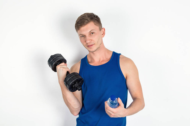 Handsome young man in blue shirt holding a dumbbell and a water bottle. White background - Photo, Image