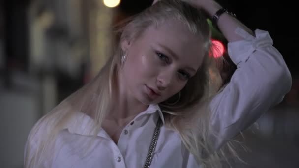 Ładna Blondynka modelka stwarzająca Dotykanie długich włosów w nocnym mieście - Materiał filmowy, wideo
