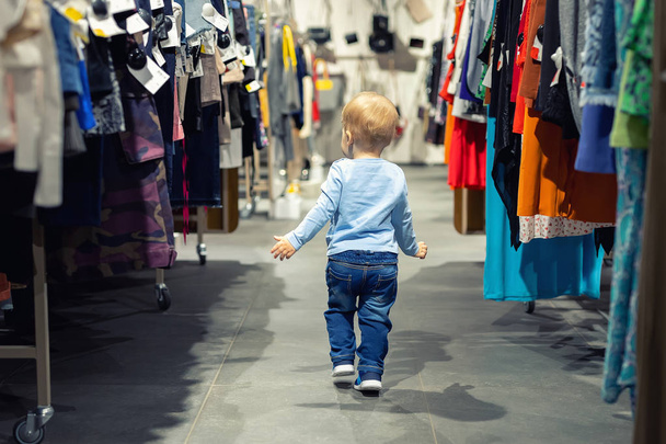 Cute kaukaski blond chłopiec maluch chodzenie sam w sklepie ubrania między Rack z wieszaki. Dziecko odkrywa świat zakupów dorosłych. Baby zgubić się w dużym hipermarketu centrum handlowe - Zdjęcie, obraz