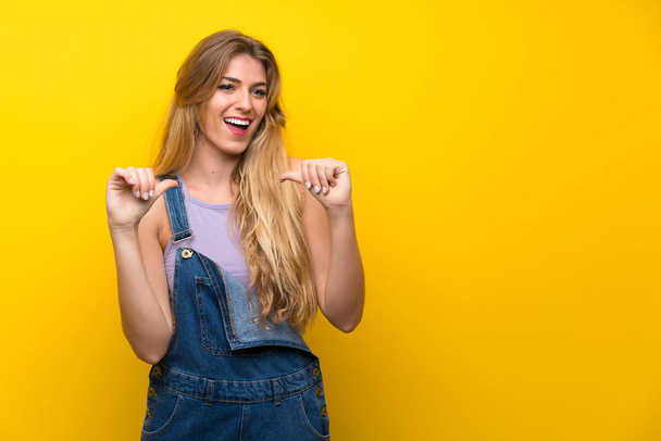 Young blonde woman with overalls over isolated yellow background proud and self-satisfied - Photo, Image