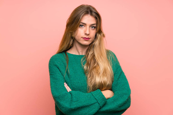 Young blonde woman with green sweater over pink wall keeping arms crossed - Photo, Image