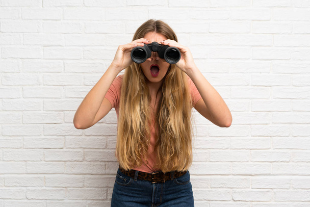 Young blonde woman over white brick wall with black binoculars - Photo, Image
