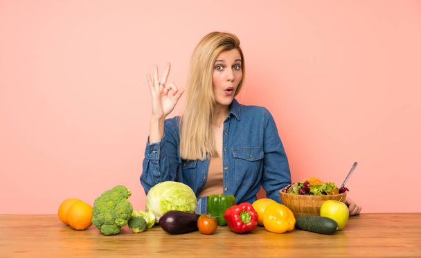 Jonge blonde vrouw met veel groenten verrast en tonen ok teken - Foto, afbeelding