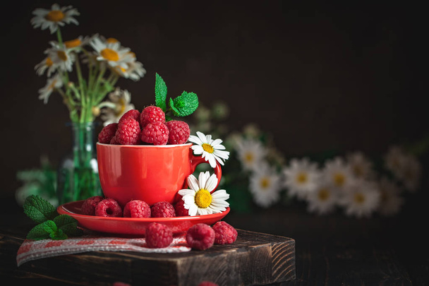 Raspberry in a red cup with chamomile and leaves on a dark background. Summer and healthy food concept. Background with copy space. Selective focus. - Foto, imagen