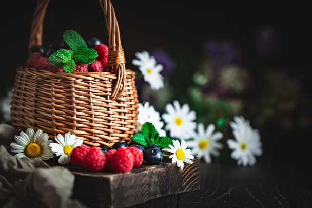 Raspberries and blueberries in a basket with chamomile and leaves on a dark background. Summer and healthy food concept. Background with copy space. Selective focus. - Photo, image