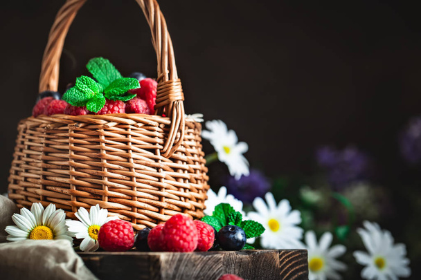 Raspberries and blueberries in a basket with chamomile and leaves on a dark background. Summer and healthy food concept. Background with copy space. Selective focus. - Foto, afbeelding