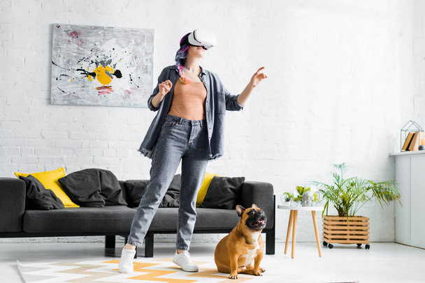girl with colorful hair and VR headset standing near dog in living room - Foto, imagen
