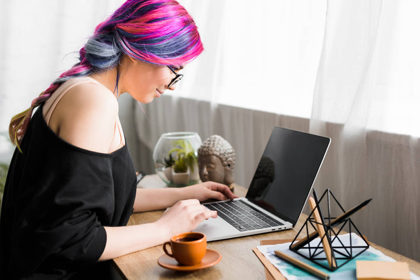 Widok z boku dziewczyny z kolorowymi włosami siedzi na biurku i za pomocą laptopa  - Zdjęcie, obraz