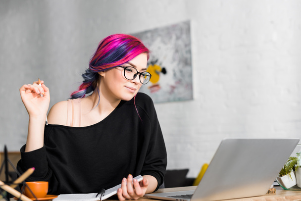 girl with colorful hair sitting at desk and making notes in living room - Foto, Imagen
