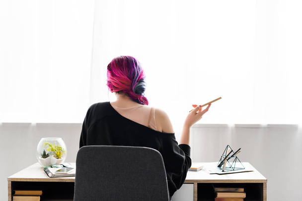 visão traseira da menina hipster segurando lápis e sentado à mesa na sala de estar
 - Foto, Imagem