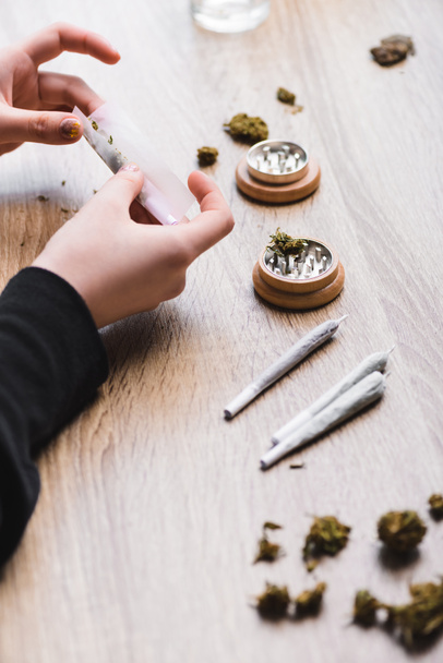 cropped view of woman making joint from weed and sitting at table with herb grinder  - Fotoğraf, Görsel