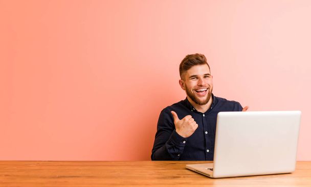 Молодий чоловік, який працює зі своїм ноутбуком, піднімає обидва великі пальці вгору, посміхаючись і впевнено
. - Фото, зображення