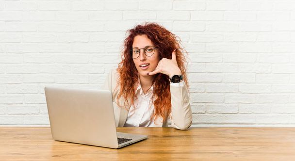 Młoda ruda kręcona kobieta pracująca z laptopem pokazująca gest rozmowy przez telefon komórkowy palcami. - Zdjęcie, obraz