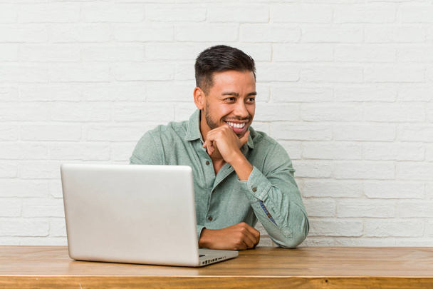 Jeune homme philippin assis travaillant avec son ordinateur portable garde les mains sous le menton, regarde joyeusement de côté
. - Photo, image
