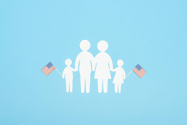 青い背景に木製の棒に装飾的なアメリカの旗を保持する白い紙カット家族のトップビュー - 写真・画像