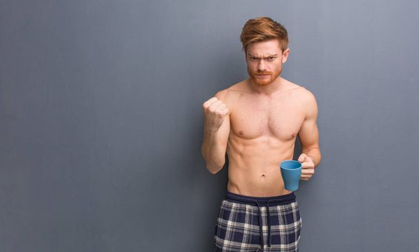 Young shirtless redhead man showing fist to front, angry expression. He is holding a coffee mug. - Photo, image