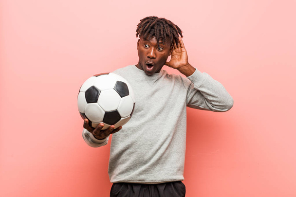 Joven fitness hombre negro sosteniendo una pelota de fútbol tratando de escuchar un chisme
. - Foto, Imagen