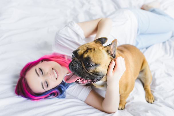 selective focus of french bulldog and happy girl with colorful hair laying with in bed  - Foto, immagini