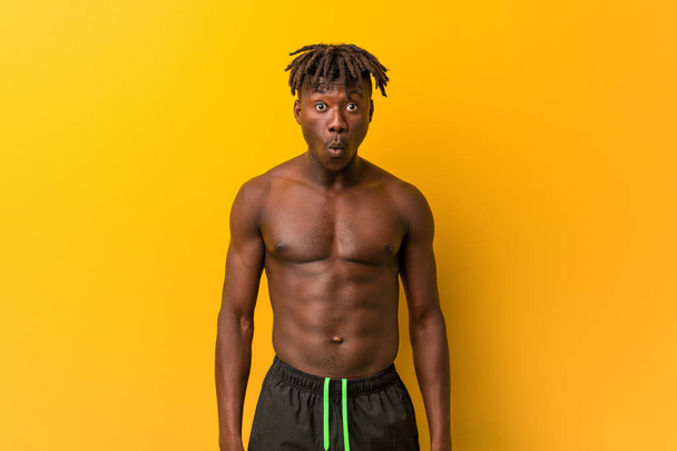 Young black man shirtless wearing swimsuit shrugs shoulders and open eyes confused. - 写真・画像