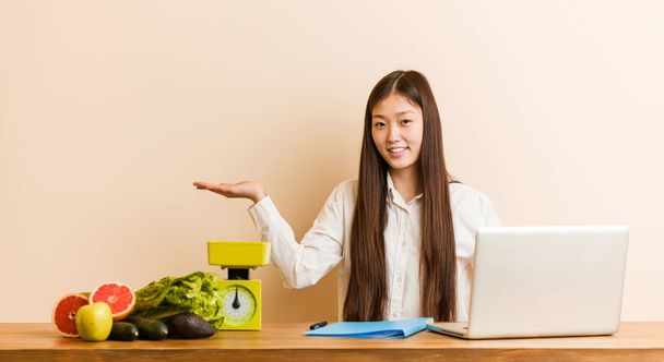 Jovem nutricionista mulher chinesa trabalhando com seu laptop mostrando um espaço de cópia em uma palma e segurando outra mão na cintura
. - Foto, Imagem