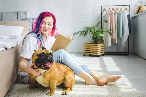  beautiful girl with colorful hair holding book, smiling and sitting on floor near French bulldog - Zdjęcie, obraz