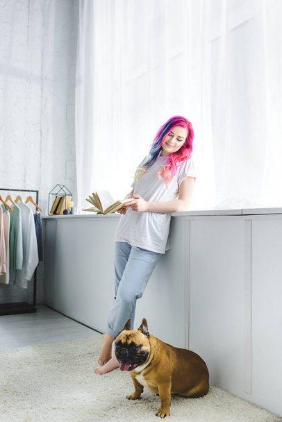 beautiful girl with colorful hair holding book and looking at French bulldog - Foto, afbeelding