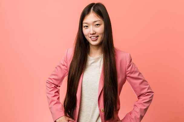 Молодая деловая китаянка в розовом костюме уверенно держит руки на бедрах
. - Фото, изображение