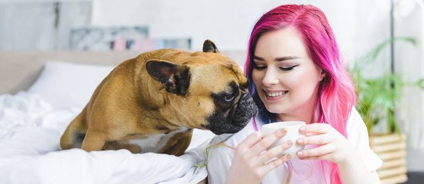 panoramic shot of happy girl with colorful hair holding cup with coffee and cute bulldog sniffing coffee - Photo, Image