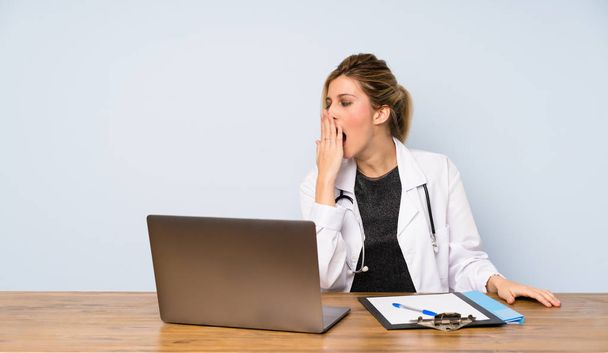 Blonde doctor woman yawning and covering wide open mouth with hand - Фото, изображение