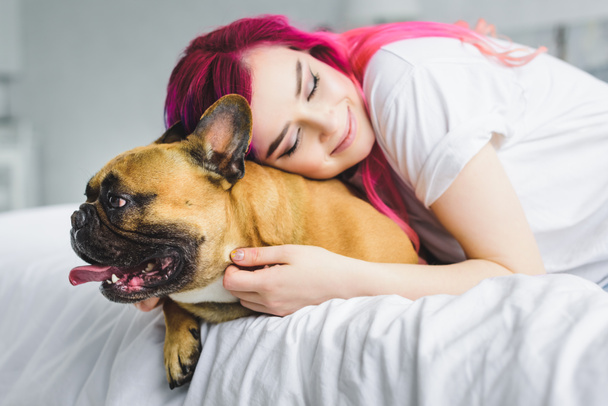  beautiful girl with colorful hair hugging cute bulldog while laying in bed  - Foto, Bild