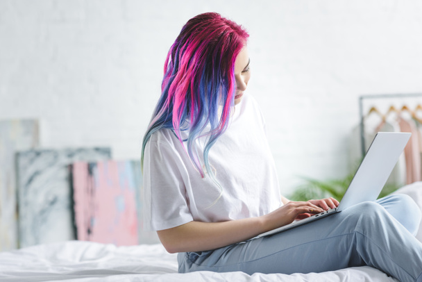 menina com cabelo colorido sentado na cama e usando laptop
  - Foto, Imagem