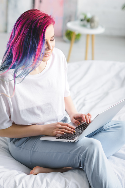 приваблива дівчина з барвистим волоссям, сидячи в ліжку, посміхаючись і використовуючи ноутбук
  - Фото, зображення
