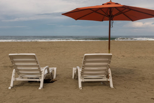 コスタリカのさびれたビーチのオレンジ色のビーチ傘の下に2つの空のプラスチック製の白い椅子 - 写真・画像